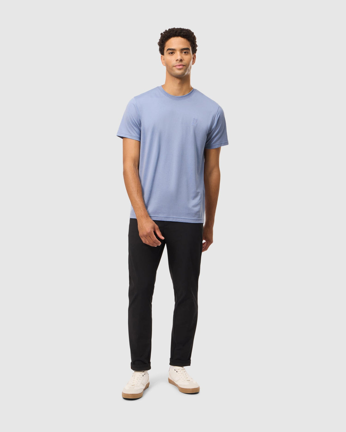 Against a plain background, a person wears the Psycho Bunny MENS JAMES PREMIUM CREW NECK TEE in blue, crafted from high-quality Pima cotton, paired with black pants and white sneakers.