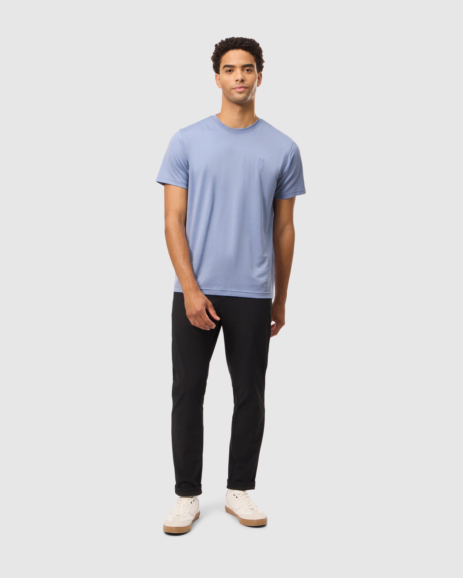 Against a plain background, a person wears the Psycho Bunny MENS JAMES PREMIUM CREW NECK TEE in blue, crafted from high-quality Pima cotton, paired with black pants and white sneakers.