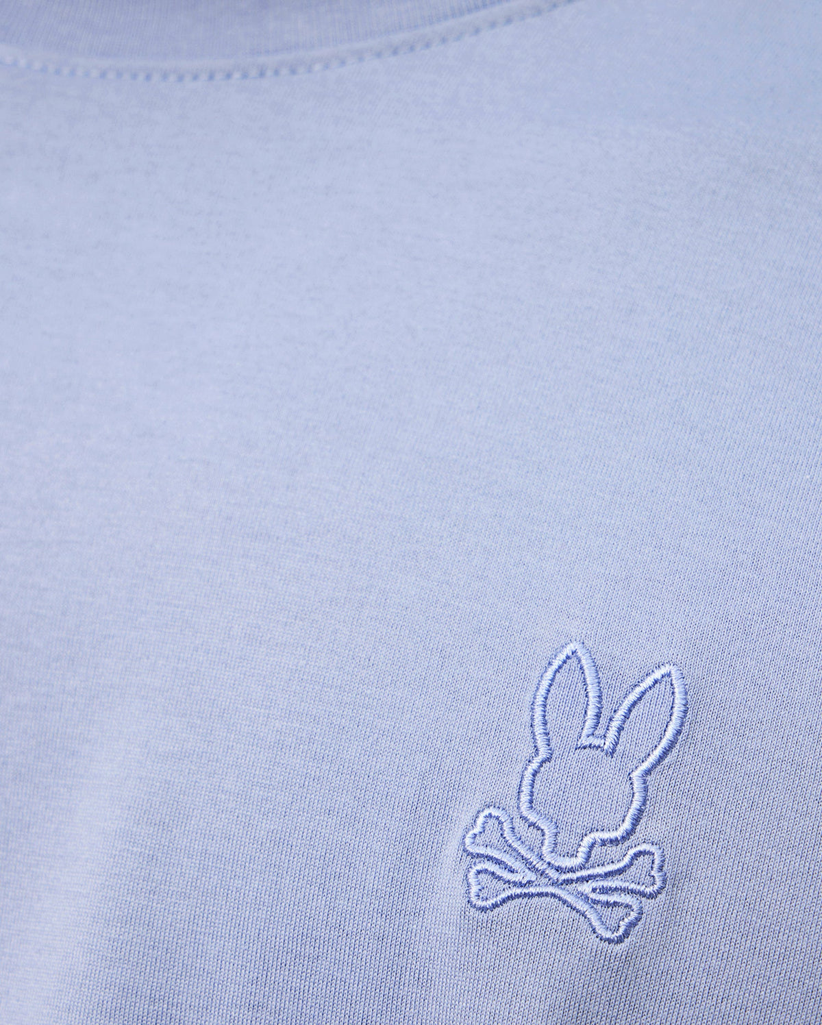 A close-up of the high-quality light blue MENS JAMES PREMIUM CREW NECK TEE by Psycho Bunny, made from soft Pima cotton and featuring a small white embroidered logo of a bunny head above crossed bones near the chest.