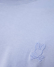 A close-up of the high-quality light blue MENS JAMES PREMIUM CREW NECK TEE by Psycho Bunny, made from soft Pima cotton and featuring a small white embroidered logo of a bunny head above crossed bones near the chest.