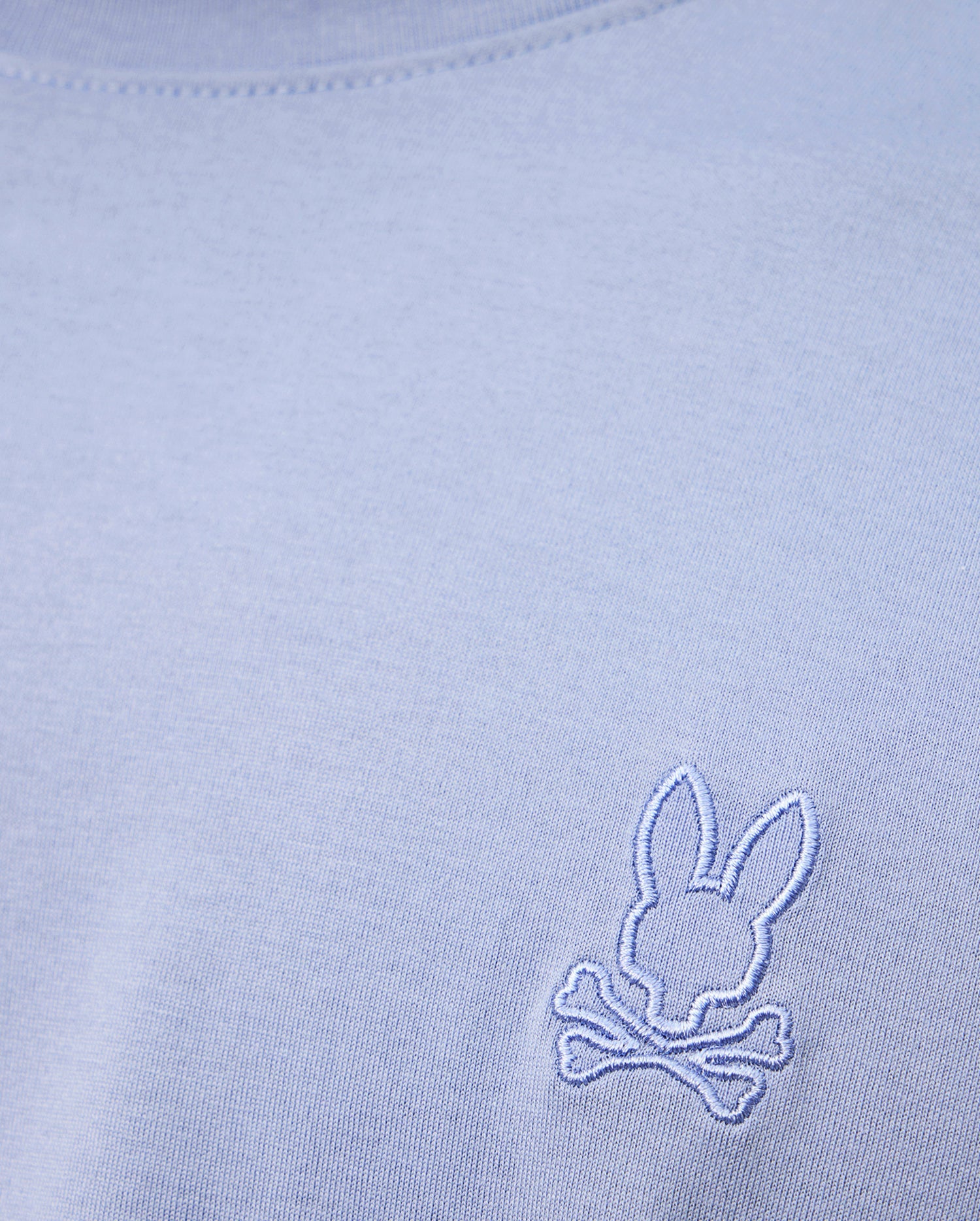 A close-up of the high-quality light blue MENS JAMES PREMIUM CREW NECK TEE by Psycho Bunny, made from soft Pima cotton and featuring a small white embroidered logo of a bunny head above crossed bones near the chest.