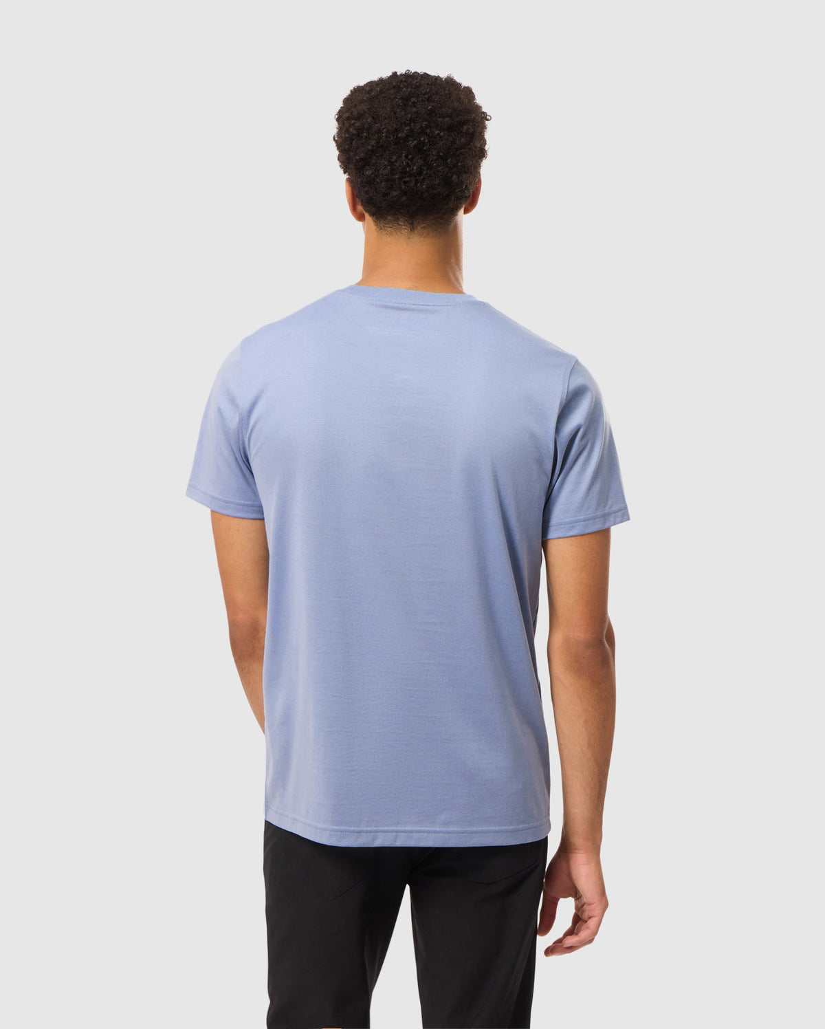 A person dressed in a Psycho Bunny Mens James Premium Crew Neck Tee, which is crafted from premium quality Pima cotton in light blue with embroidery detail, and black pants is standing with their back to the camera against a plain white background.