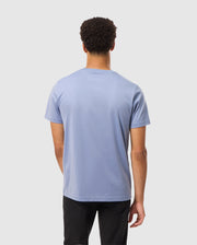 A person dressed in a Psycho Bunny Mens James Premium Crew Neck Tee, which is crafted from premium quality Pima cotton in light blue with embroidery detail, and black pants is standing with their back to the camera against a plain white background.