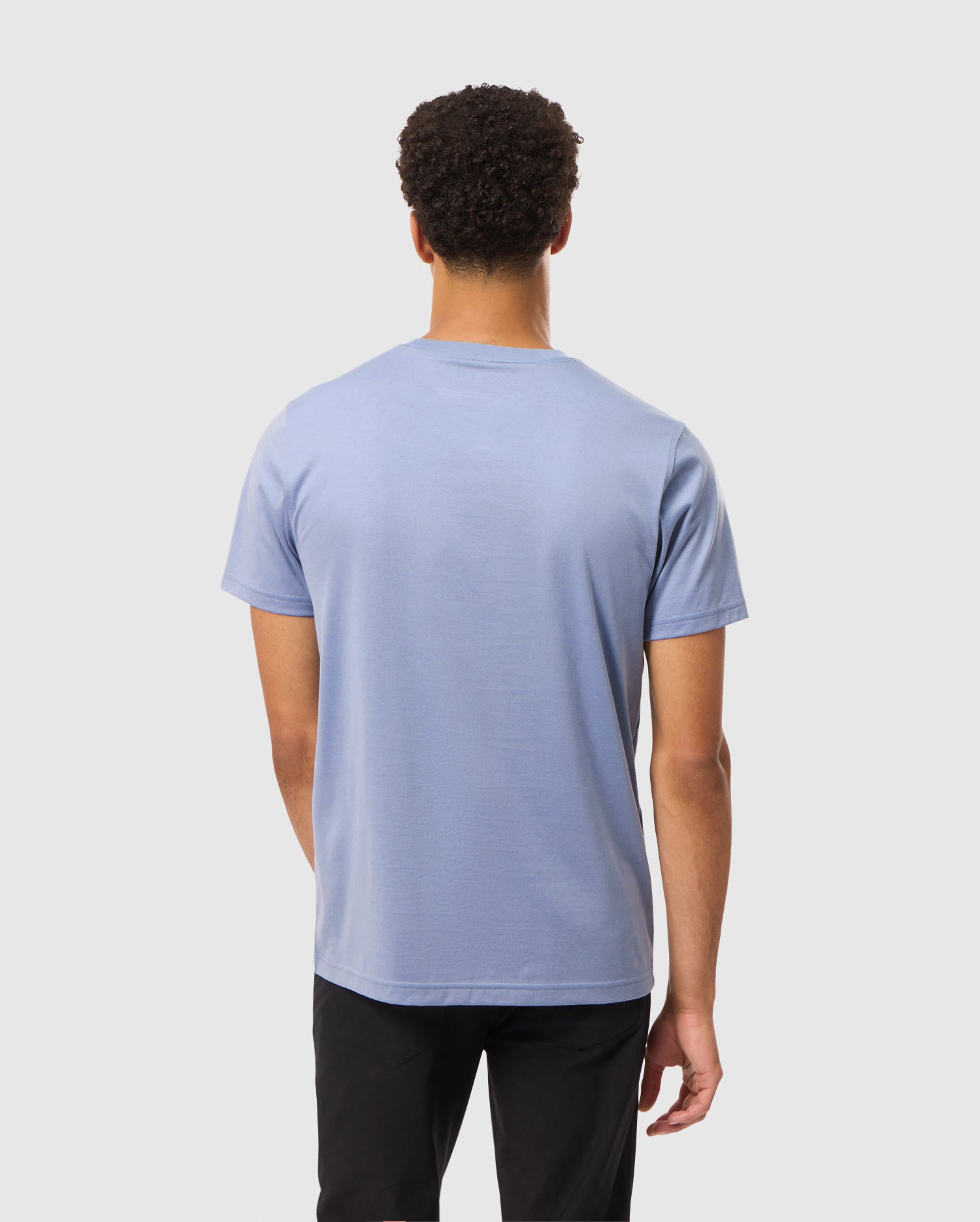 A person dressed in a Psycho Bunny Mens James Premium Crew Neck Tee, which is crafted from premium quality Pima cotton in light blue with embroidery detail, and black pants is standing with their back to the camera against a plain white background.