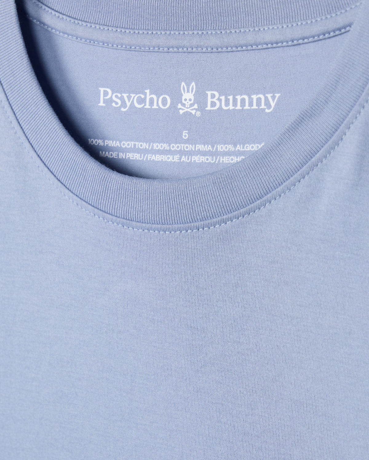 Close-up of a blue t-shirt highlighting the Psycho Bunny logo and brand name inside the neckline. The label proudly declares that the MENS JAMES PREMIUM CREW NECK TEE - B6U952D200 is crafted from 100% premium Pima cotton and manufactured in Peru, embodying the premium quality that defines this embroidered tee.