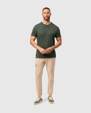 A person wearing a stylish Psycho Bunny MENS JAMES PREMIUM CREW NECK TEE in dark green, paired with beige pants and green sneakers, stands against a light gray background, holding their hands together in front.