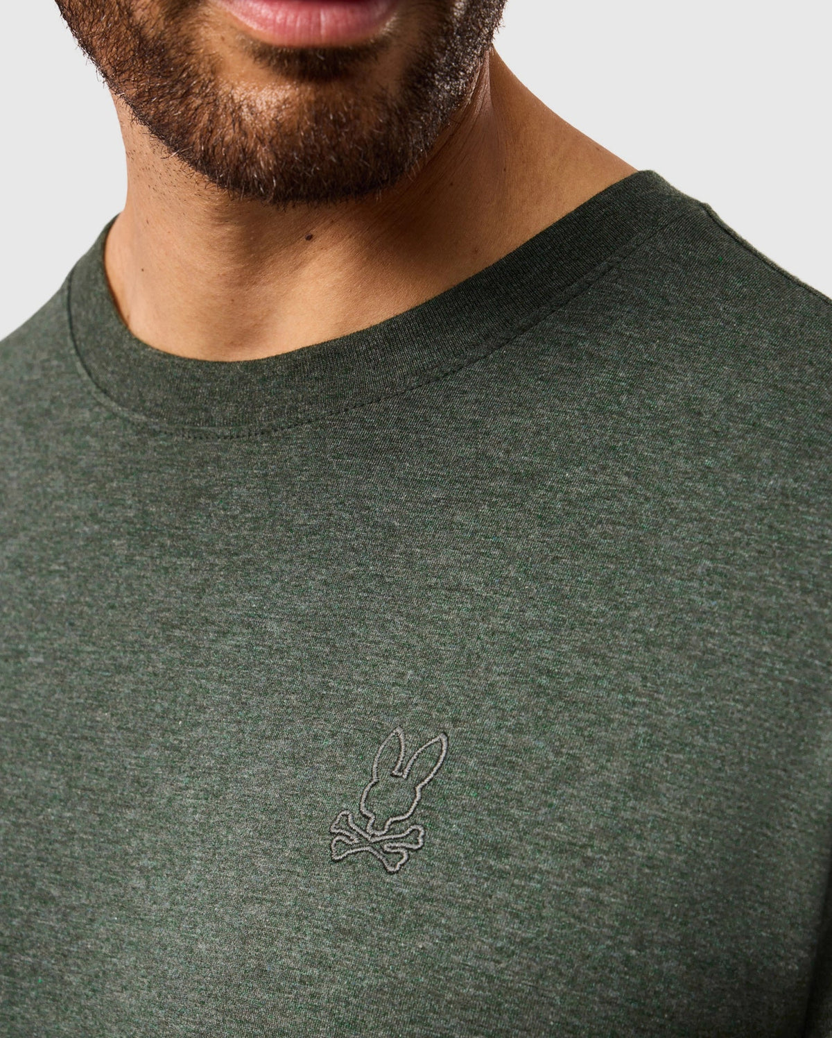 A person wearing the Psycho Bunny MENS JAMES PREMIUM CREW NECK TEE - B6U952D200, which is a stylish dark green T-shirt made from premium Pima cotton. The shirt features a small tonal outline bunny logo embroidered on the chest, showcasing a rabbit's head over crossed bones. The image highlights the chest area with the person's face partially visible.