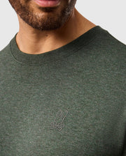 A person wearing the Psycho Bunny MENS JAMES PREMIUM CREW NECK TEE - B6U952D200, which is a stylish dark green T-shirt made from premium Pima cotton. The shirt features a small tonal outline bunny logo embroidered on the chest, showcasing a rabbit's head over crossed bones. The image highlights the chest area with the person's face partially visible.