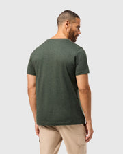 A man with short hair is standing with his back turned, wearing a Psycho Bunny MENS JAMES PREMIUM CREW NECK TEE - B6U952D200 in a stylish dark green made of premium Pima cotton, along with light beige pants. The shirt showcases a tonal outline bunny embroidery. The background is a simple light gray.