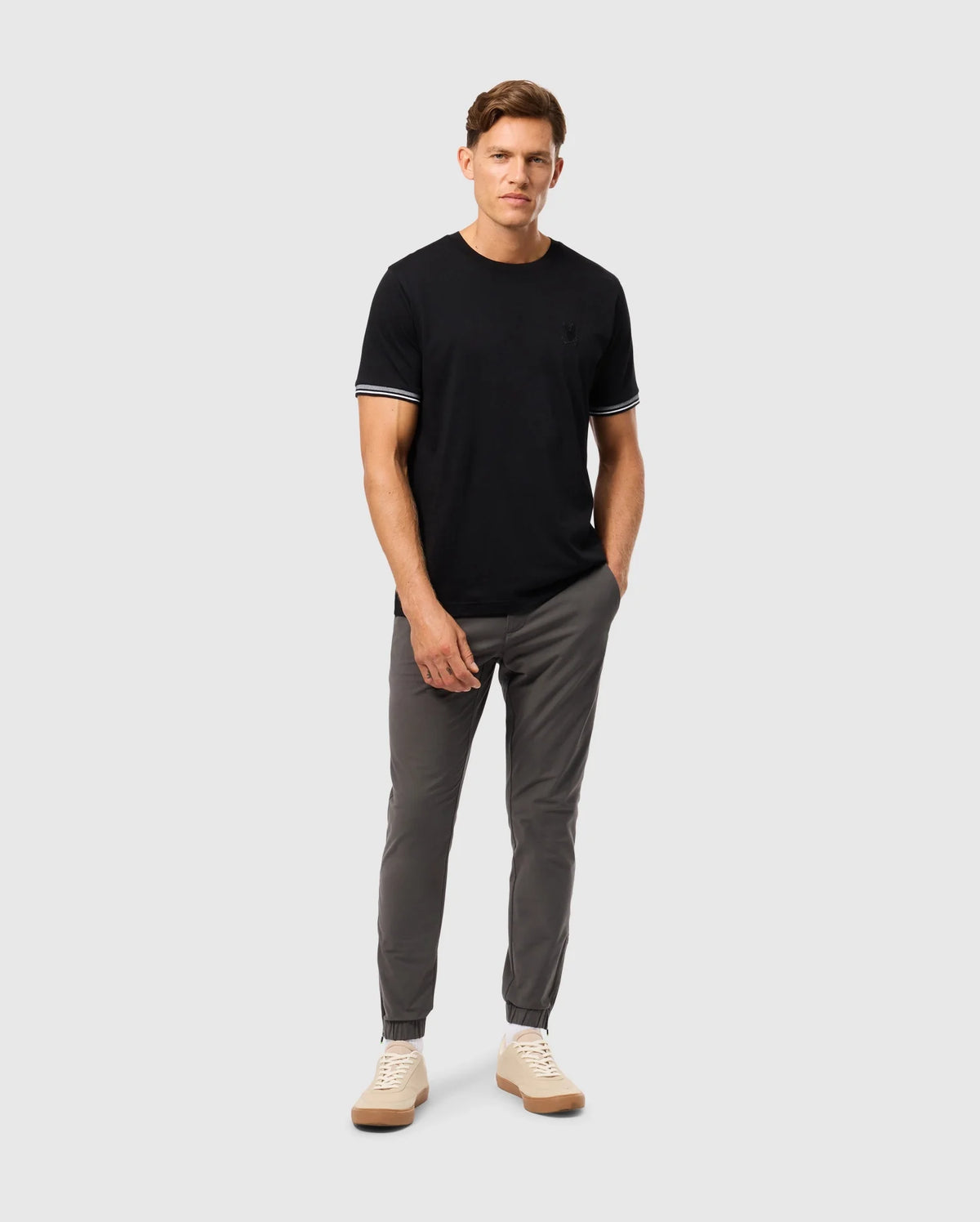 A person stands against a light gray background wearing a stylish Psycho Bunny MENS MALTON FASHION TEE, featuring white trim on the sleeves, paired with gray pants and beige sneakers. They have their left hand in their pocket and are looking forward.
