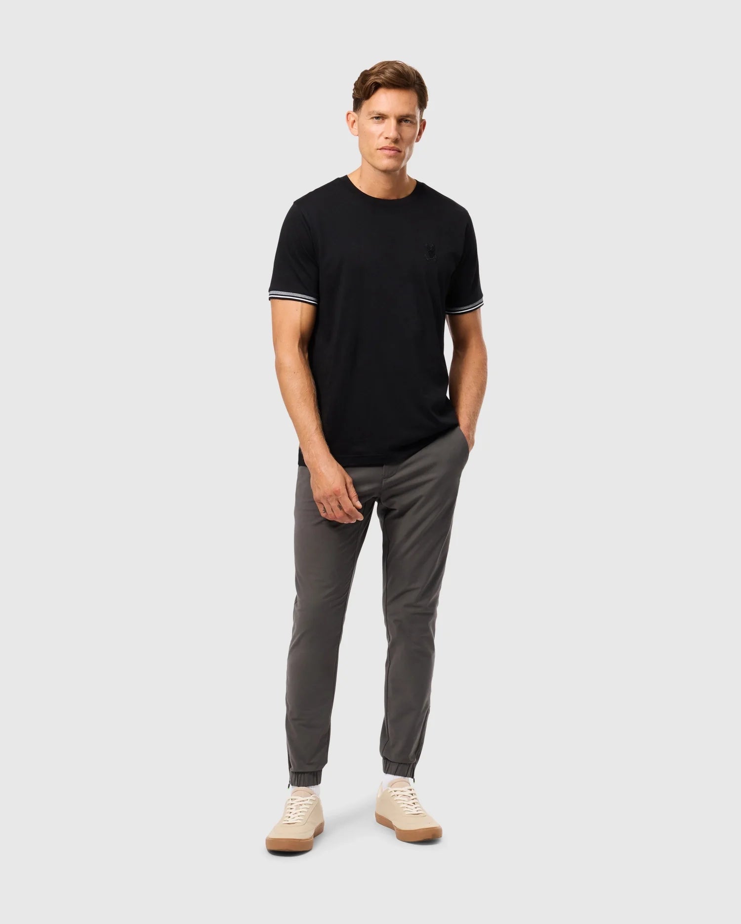 A person stands against a light gray background wearing a stylish Psycho Bunny MENS MALTON FASHION TEE, featuring white trim on the sleeves, paired with gray pants and beige sneakers. They have their left hand in their pocket and are looking forward.