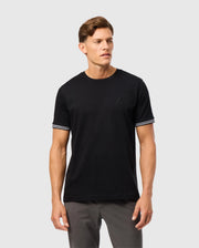 A person wearing the Psycho Bunny MENS MALTON FASHION TEE - B6U966D200, a stylish cotton tee with white striped trim on the sleeves, and gray pants stands against a light gray background. The men's fashion tee features a subtle logo on the chest, blending comfort with modern style.