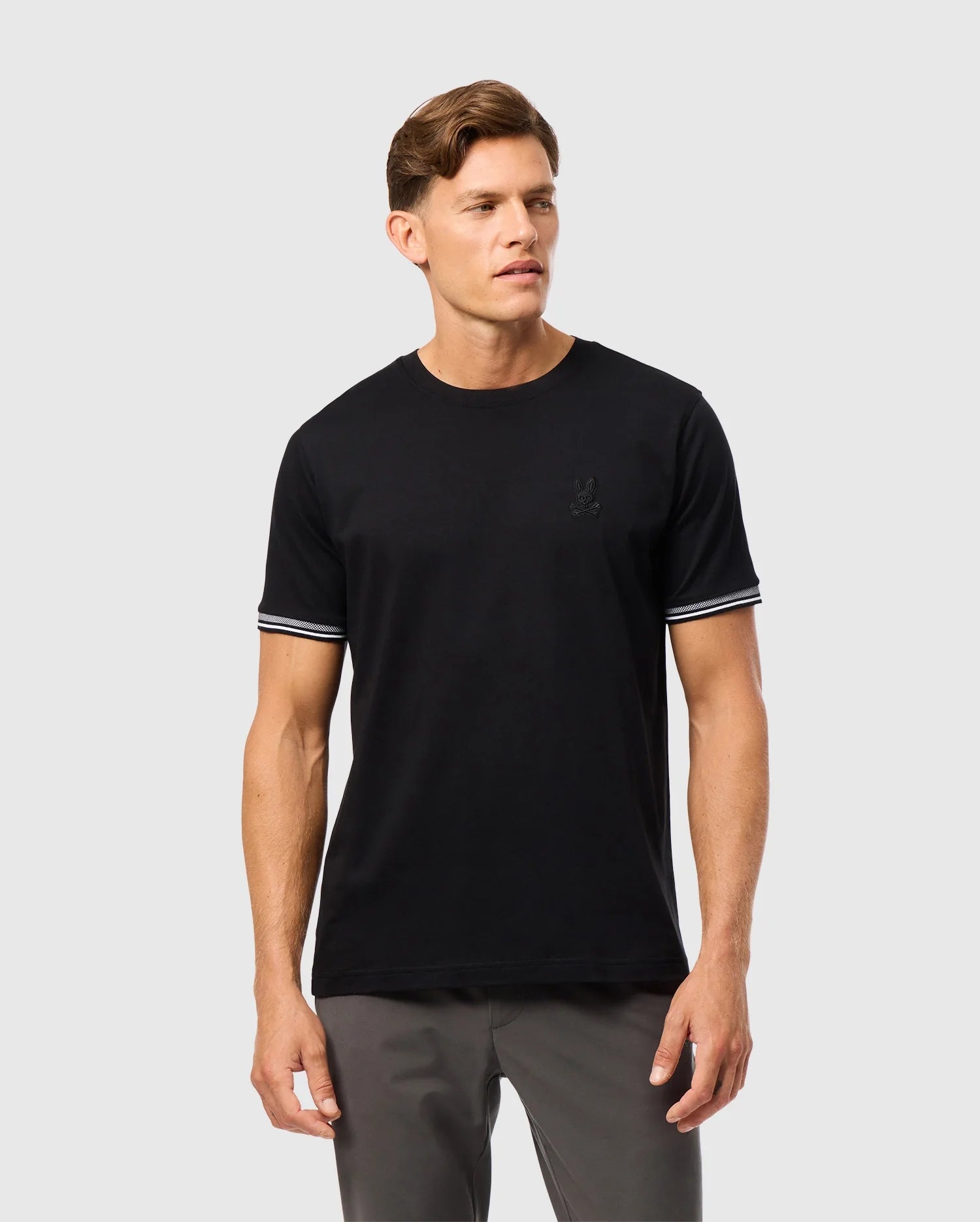 A person wearing the Psycho Bunny MENS MALTON FASHION TEE - B6U966D200, a stylish cotton tee with white striped trim on the sleeves, and gray pants stands against a light gray background. The men's fashion tee features a subtle logo on the chest, blending comfort with modern style.