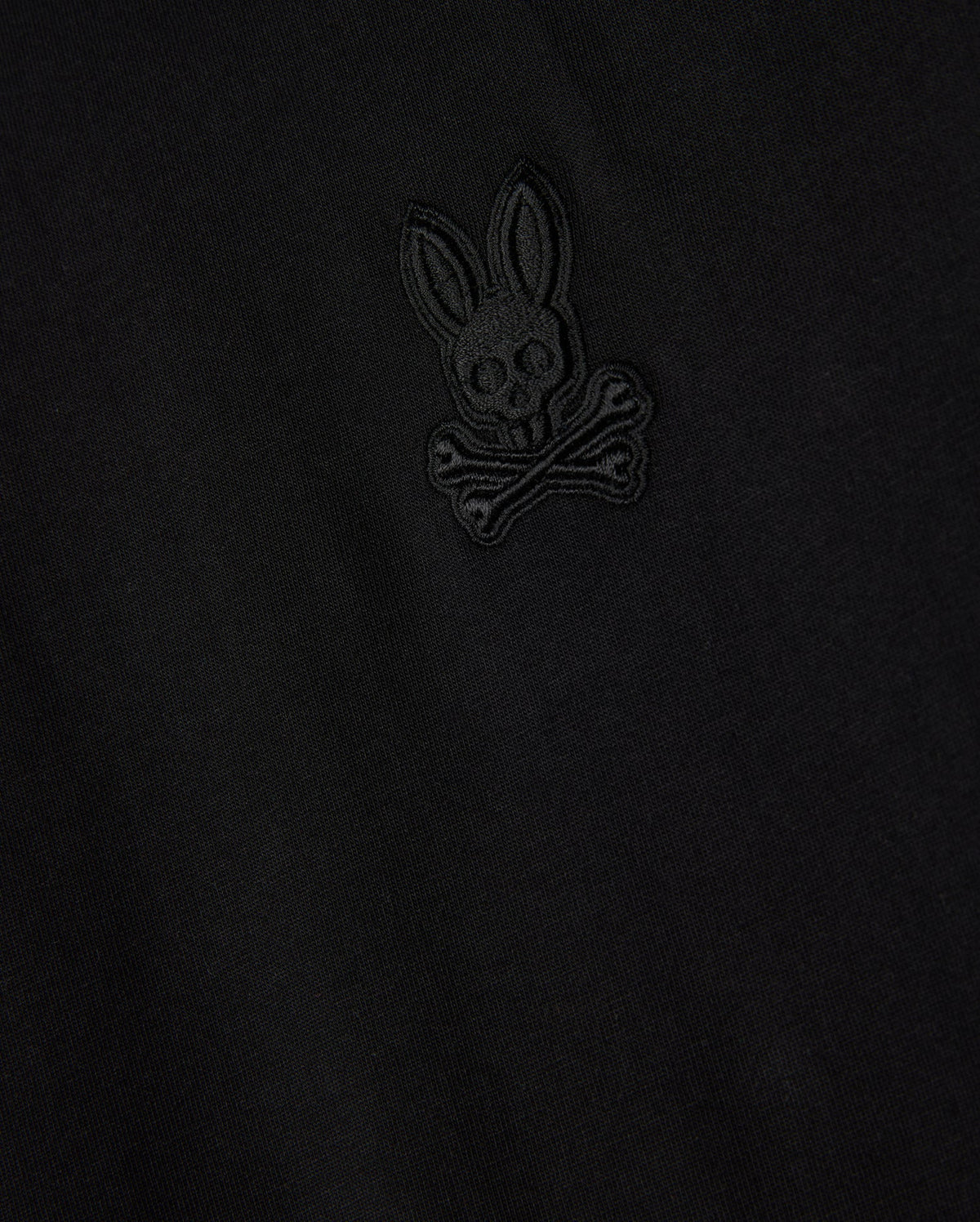 Close-up of the Psycho Bunny MENS MALTON FASHION TEE - B6U966D200, highlighting its stylish cotton fabric in black, adorned with an embroidered emblem resembling a skull and crossbones motif, featuring a rabbit head with long ears above crossed bones. The design is intricately stitched in subtle black thread, marking it as a standout piece in men's fashion.