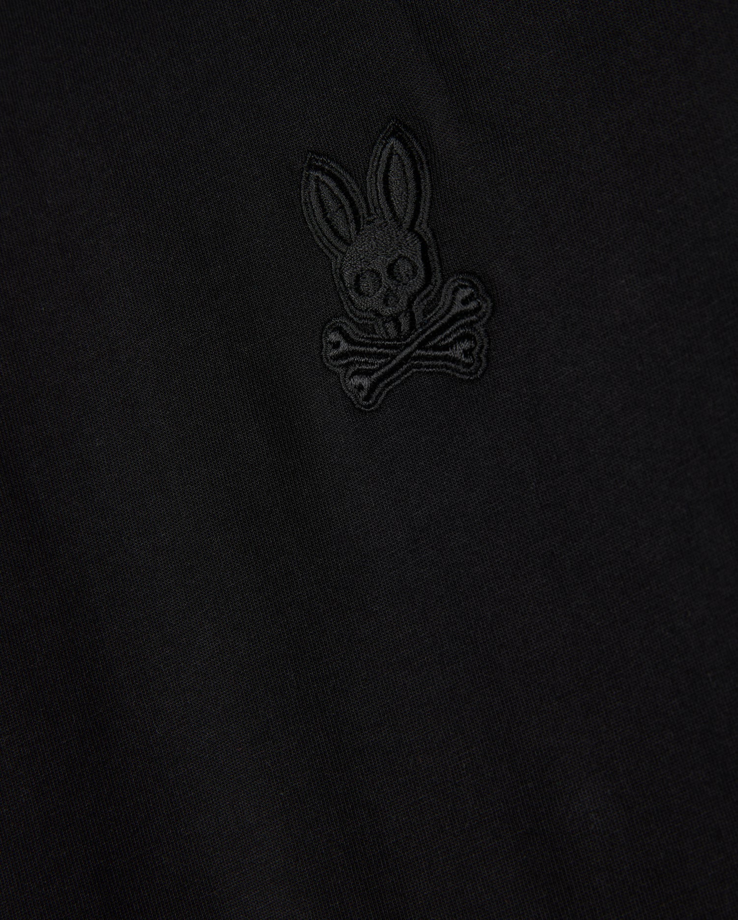 Close-up of the Psycho Bunny MENS MALTON FASHION TEE - B6U966D200, highlighting its stylish cotton fabric in black, adorned with an embroidered emblem resembling a skull and crossbones motif, featuring a rabbit head with long ears above crossed bones. The design is intricately stitched in subtle black thread, marking it as a standout piece in men's fashion.