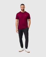 A man wearing the Psycho Bunny MENS MALTON FASHION TEE in a chic burgundy hue, complemented by dark pants and white sneakers, stands against a plain gray backdrop. With a beard and his hands resting casually in his pockets, he exudes effortless style.