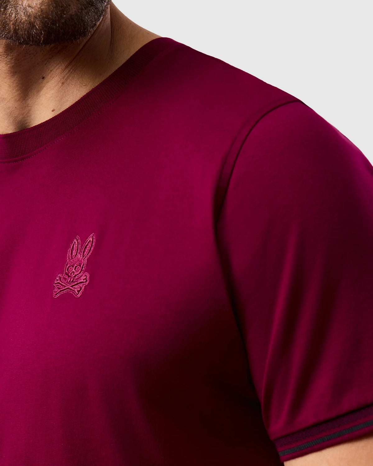 A close-up of the MENS MALTON FASHION TEE - B6U966D200 from Psycho Bunny highlights its stylish maroon color with dark trim on the collar and sleeves. Crafted from soft pima cotton, it features an embroidered logo of a cartoon rabbit. This tee is perfect for those looking for men's fashion with understated flair, set against a plain gray background.