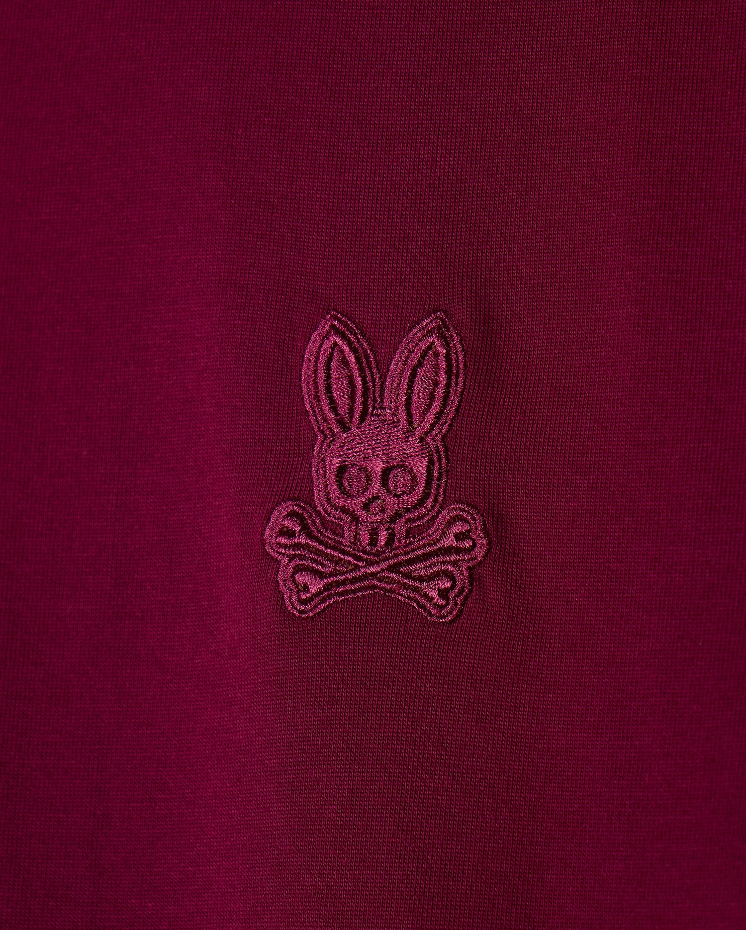 Detailed view of the maroon pima cotton fabric from the Psycho Bunny Men's Malton Fashion Tee (B6U966D200), featuring an embroidered logo of a bunny's head with crossed bones beneath. This stylish t-shirt showcases the logo stitched in a slightly darker shade of maroon for a subtle contrast, making it an excellent addition to any men's fashion tee collection.