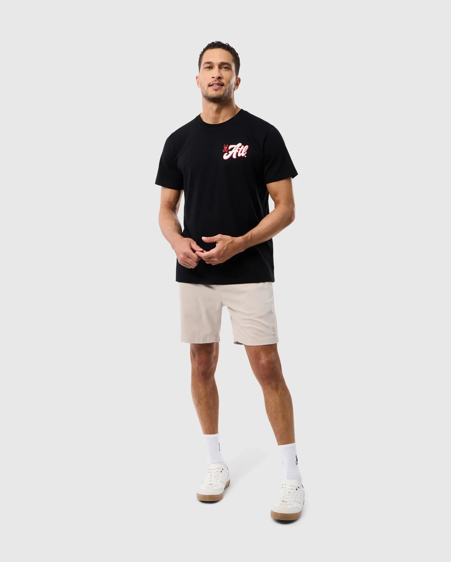 A man wearing a black Psycho Bunny ATLANTA CITY Tee with a red logo, beige shorts, and white sneakers standing against a plain white background.