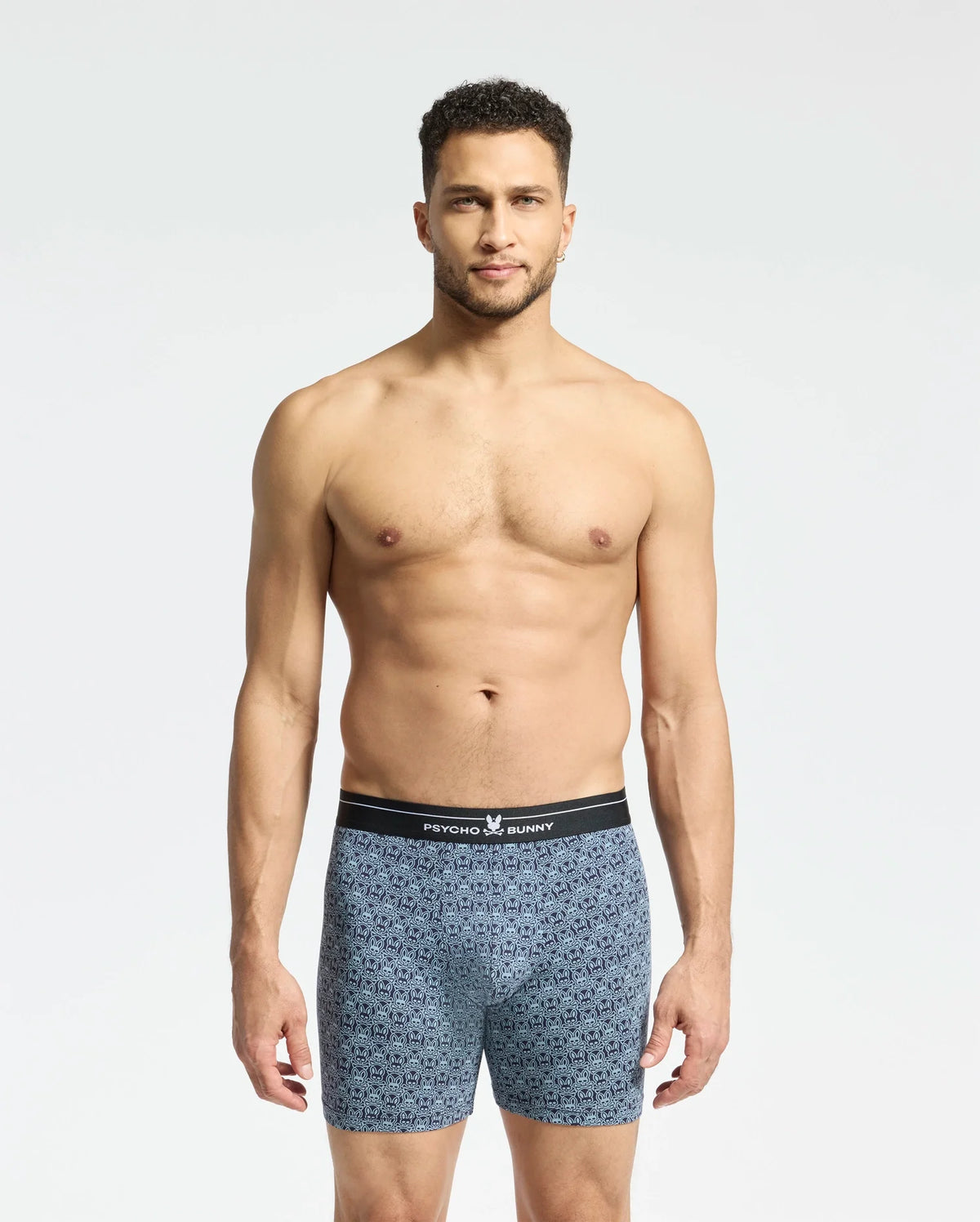 A man with short hair stands against a plain white background, wearing soft-knit patterned blue MENS 2 PACK BOXER BRIEF - B6V481F200 by Psycho Bunny. The briefs have a black waistband featuring the brand name, and his relaxed expression highlights their comfort and breathability.