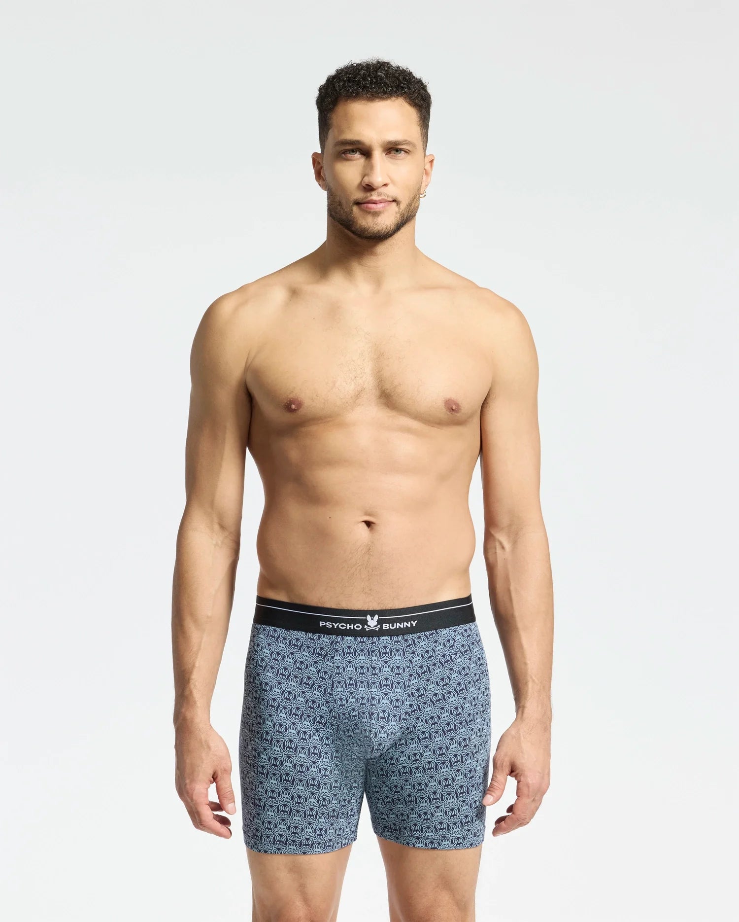 A man with short hair stands against a plain white background, wearing soft-knit patterned blue MENS 2 PACK BOXER BRIEF - B6V481F200 by Psycho Bunny. The briefs have a black waistband featuring the brand name, and his relaxed expression highlights their comfort and breathability.