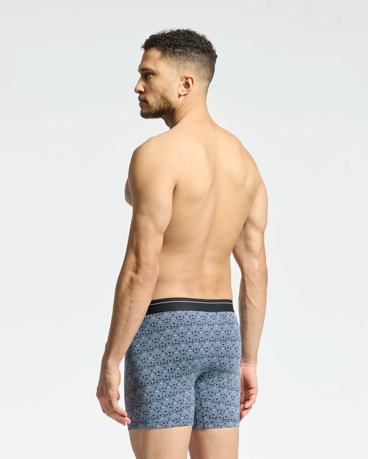 In profile, a shirtless man wearing Psycho Bunny's MENS 2 PACK BOXER BRIEF - B6V481F200 faces left, highlighting his muscular build. The breathable soft-knit fabric features intricate blue patterns against a light gray background that contrasts with his skin tone.
