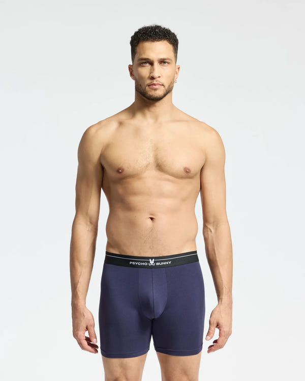 A man stands against a plain background wearing navy blue Psycho Bunny boxer briefs with a black waistband. Made from stretch Pima cotton, these breathable briefs promise supreme comfort. He looks directly at the camera with a neutral expression. Product: MENS 2 PACK BOXER BRIEF - B6V481F200.