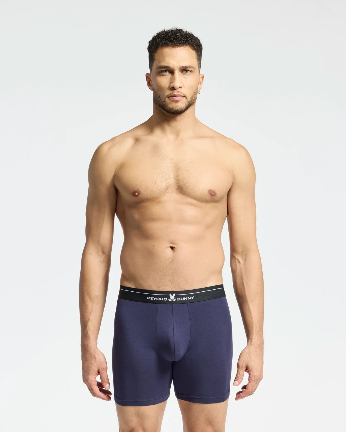 A man stands against a plain background wearing navy blue Psycho Bunny boxer briefs with a black waistband. Made from stretch Pima cotton, these breathable briefs promise supreme comfort. He looks directly at the camera with a neutral expression. Product: MENS 2 PACK BOXER BRIEF - B6V481F200.