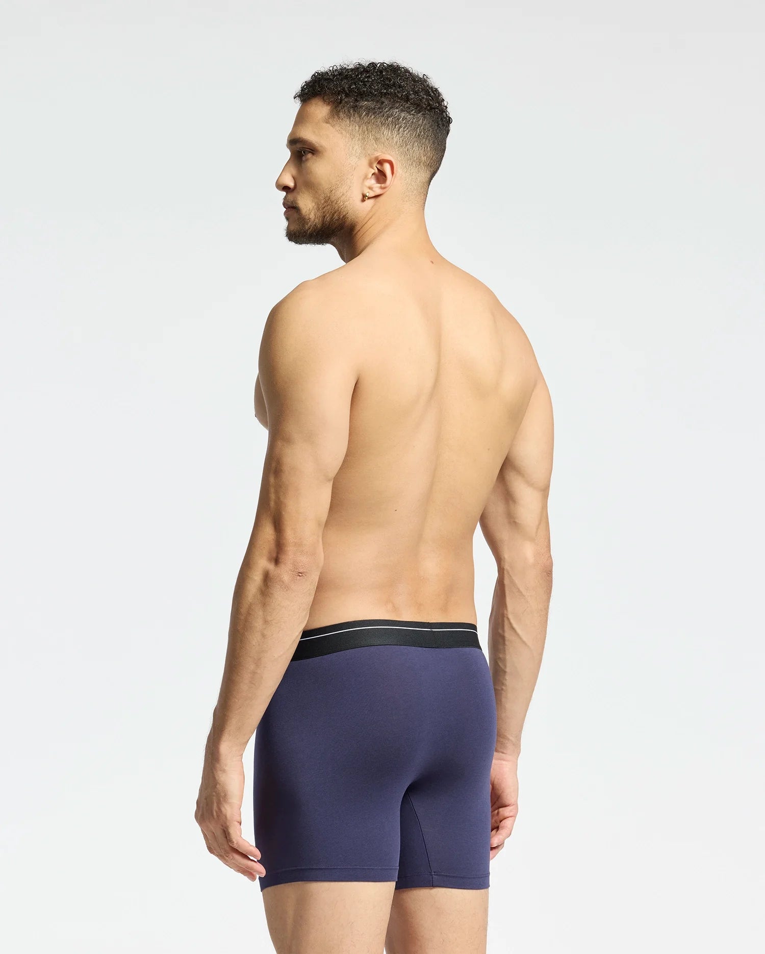 A muscular man faces away, wearing Psycho Bunny's MENS 2 PACK BOXER BRIEF - B6V481F200 in dark blue stretch Pima cotton with a black waistband. The plain light gray background highlights the breathable, soft-knit fabric.