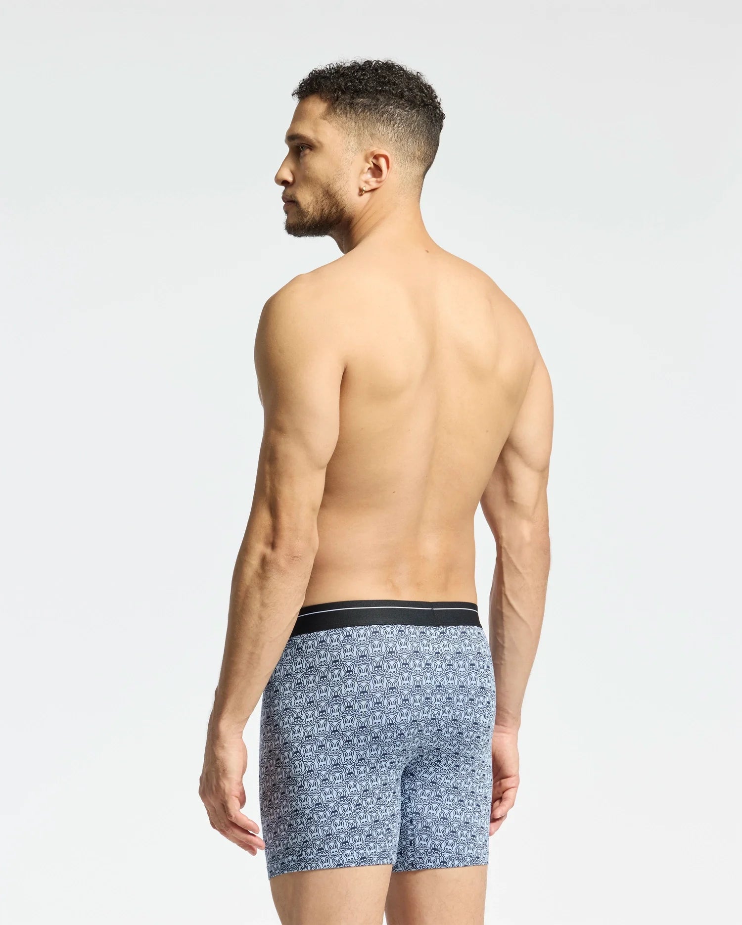 A shirtless man with short curly hair faces away, displaying Psycho Bunny's MENS 2 PACK BOXER BRIEF (B6V481F200) in a blue pattern made from a luxurious Pima cotton-Modal blend. A black waistband adds stretchy comfort against a plain white backdrop.