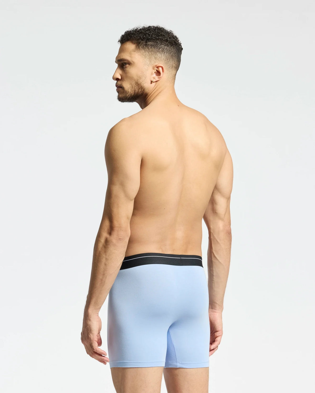 A shirtless man with curly hair stands against a plain light background, wearing Psycho Bunny's light blue boxer briefs from the MENS 2 PACK BOXER BRIEF - B6V481F200 collection, featuring breathable Stretch Pima cotton and a black waistband.