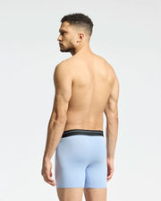 A shirtless man with curly hair stands against a plain light background, wearing Psycho Bunny's light blue boxer briefs from the MENS 2 PACK BOXER BRIEF - B6V481F200 collection, featuring breathable Stretch Pima cotton and a black waistband.