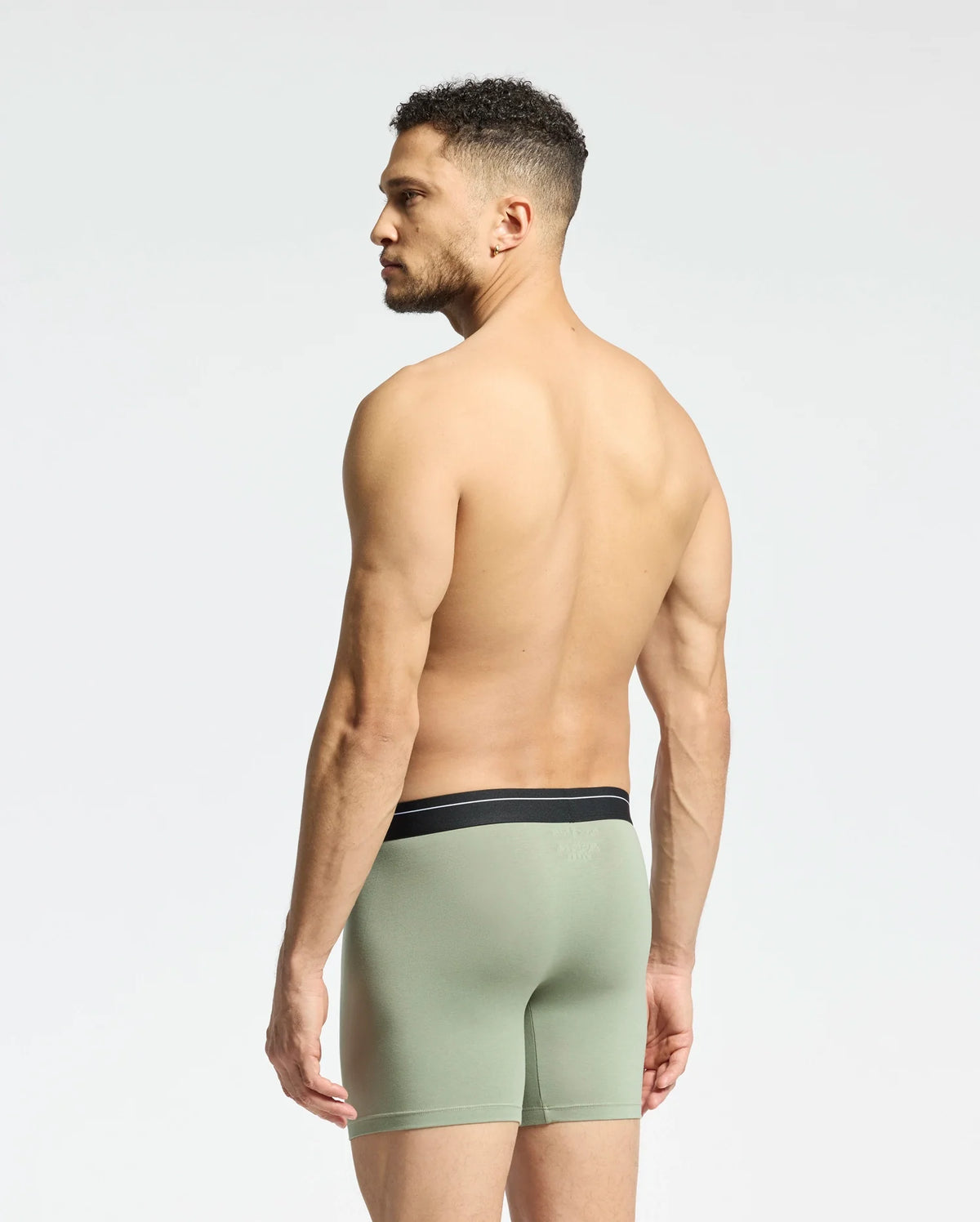 A man with short curly hair stands barefoot, partially showing his back and facing sideways, wearing Psycho Bunny's light green MENS 2 PACK BOXER BRIEF - B6V481F200. Made from a Pima cotton-Modal blend with a black waistband, these boxer briefs promise all-day comfort against a plain, light background.
