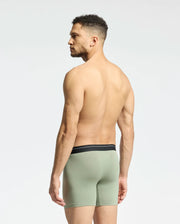 A man with short curly hair stands barefoot, partially showing his back and facing sideways, wearing Psycho Bunny's light green MENS 2 PACK BOXER BRIEF - B6V481F200. Made from a Pima cotton-Modal blend with a black waistband, these boxer briefs promise all-day comfort against a plain, light background.