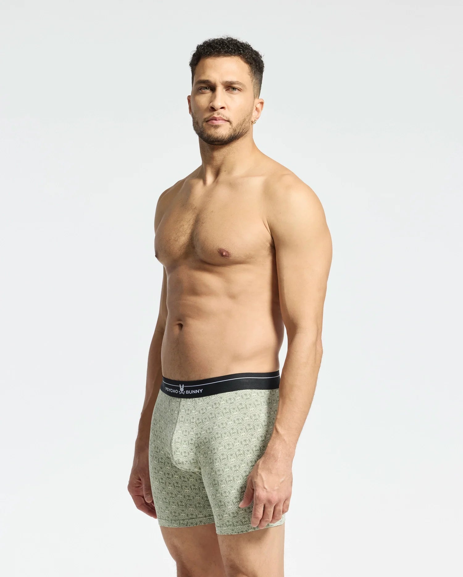 A man with short hair and a beard neutrally poses against a plain background in fitted, patterned boxers made from a Pima cotton-Modal blend. The stretchy design of Psycho Bunny's MENS 2 PACK BOXER BRIEF (B6V481F200) ensures all-day comfort with a dark waistband featuring the visible logo.
