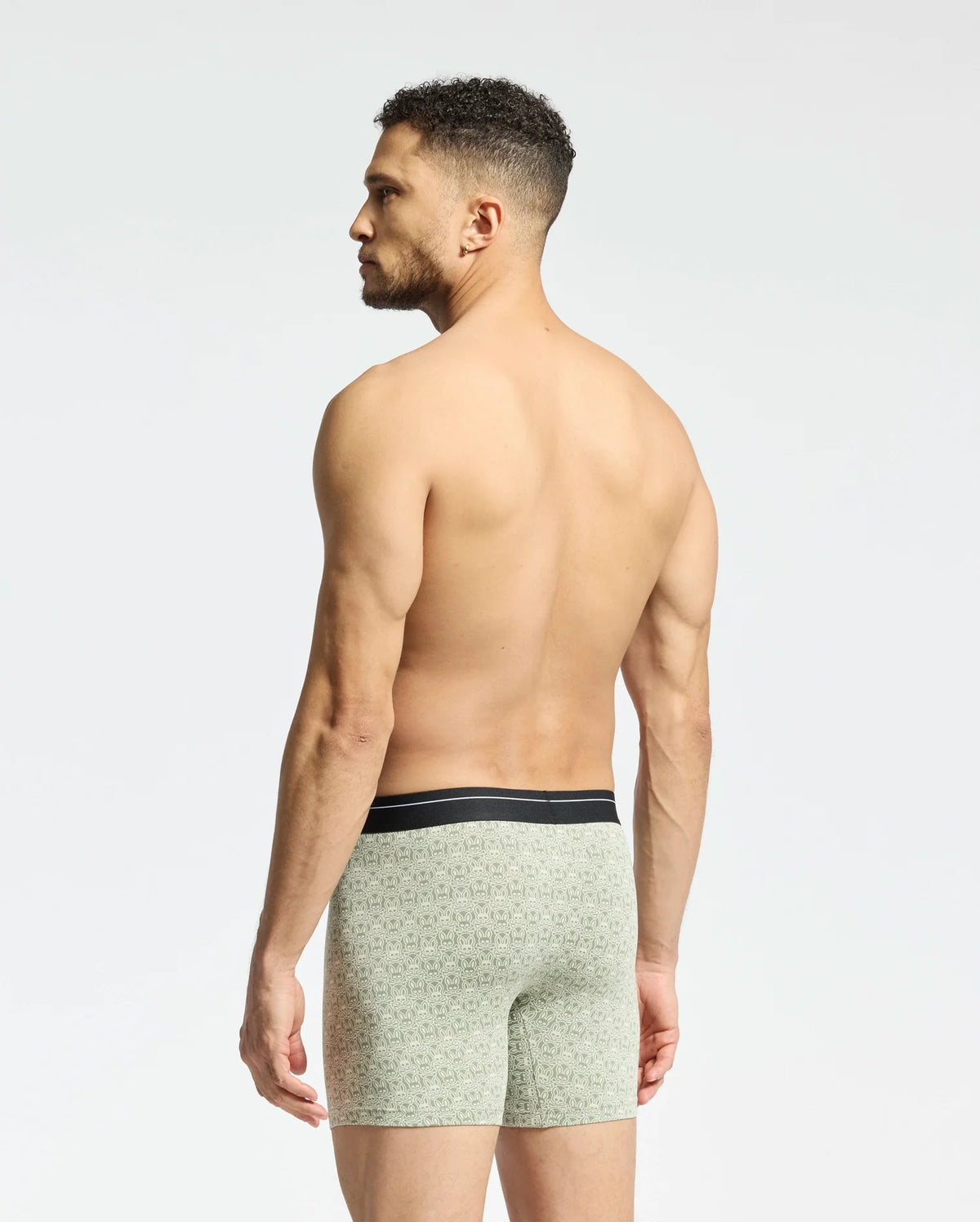 A man with short curly hair stands shirtless facing away, wearing Psycho Bunny's MENS 2 PACK BOXER BRIEF - B6V481F200 in light green with patterns and a black waistband. Made from a stretchy Pima cotton-Modal blend, they ensure all-day comfort against a plain white background.