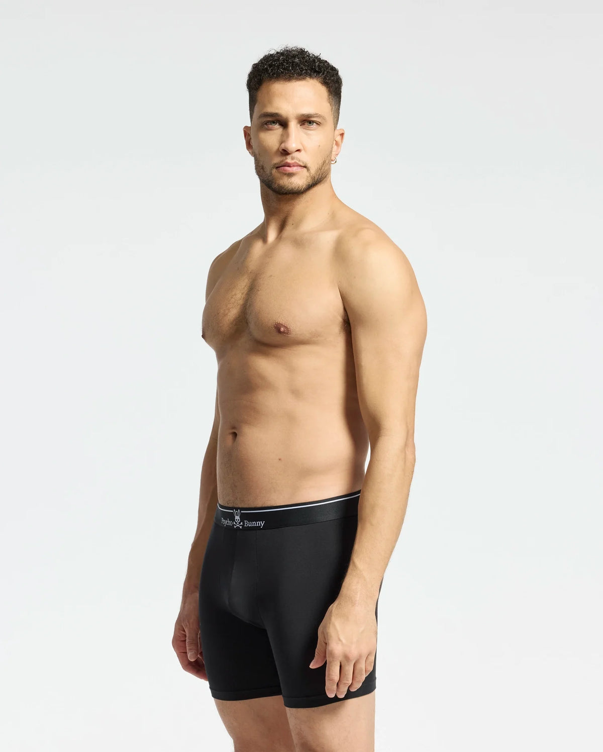 A man stands against a light gray background wearing the MENS SOLID KNIT 2 PACK BOXER BRIEF - B6V906ARCM by Psycho Bunny. He looks ahead with his arms at his sides, showcasing comfort and style in these ultrasoft modal-blend boxer briefs.