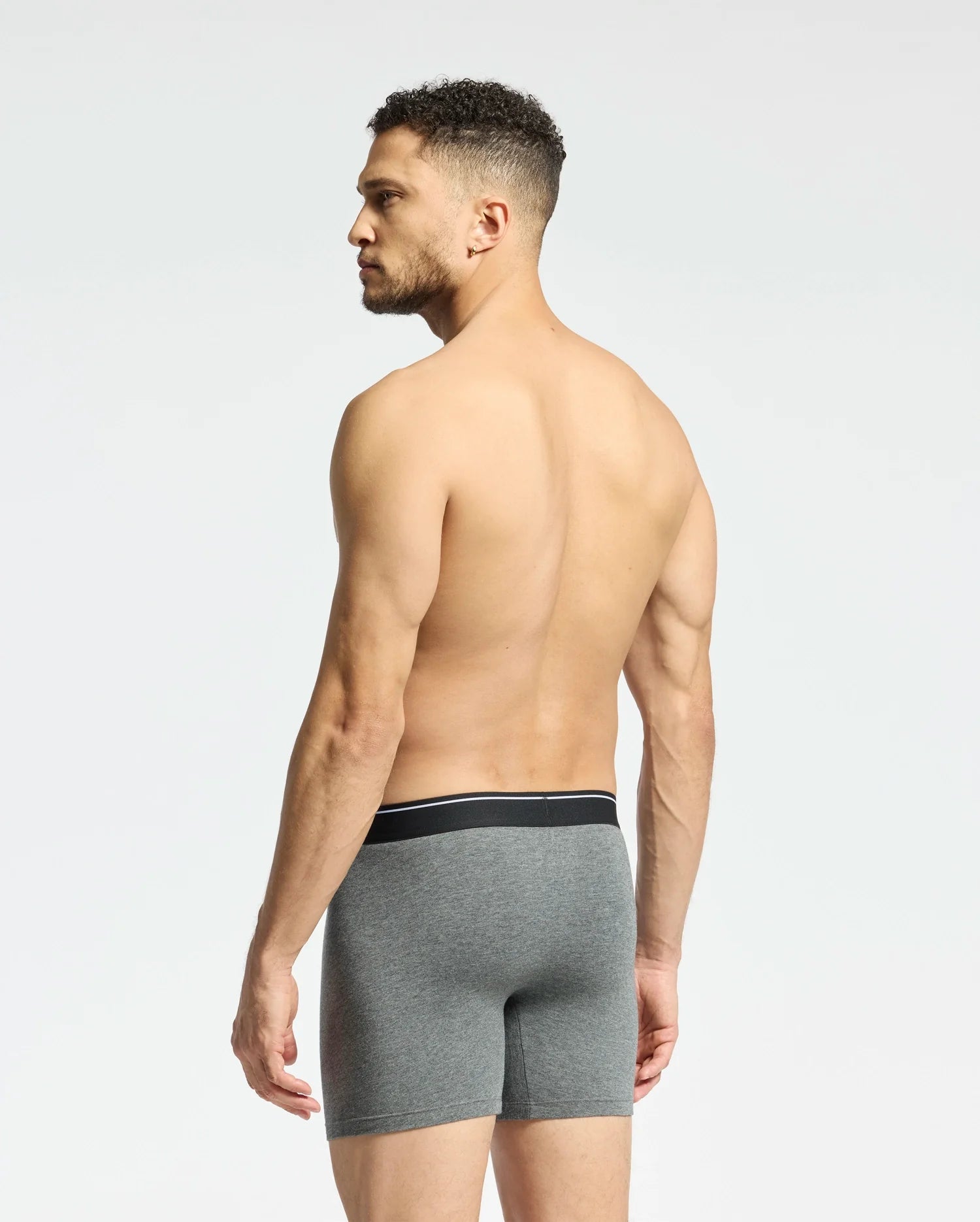 A shirtless, curly-haired man faces away from the camera, highlighting the comfort of Psycho Bunny’s gray MENS SOLID KNIT 2 PACK BOXER BRIEF - B6V906ARCM made from smooth Pima cotton with a sleek black waistband against a plain, light background.