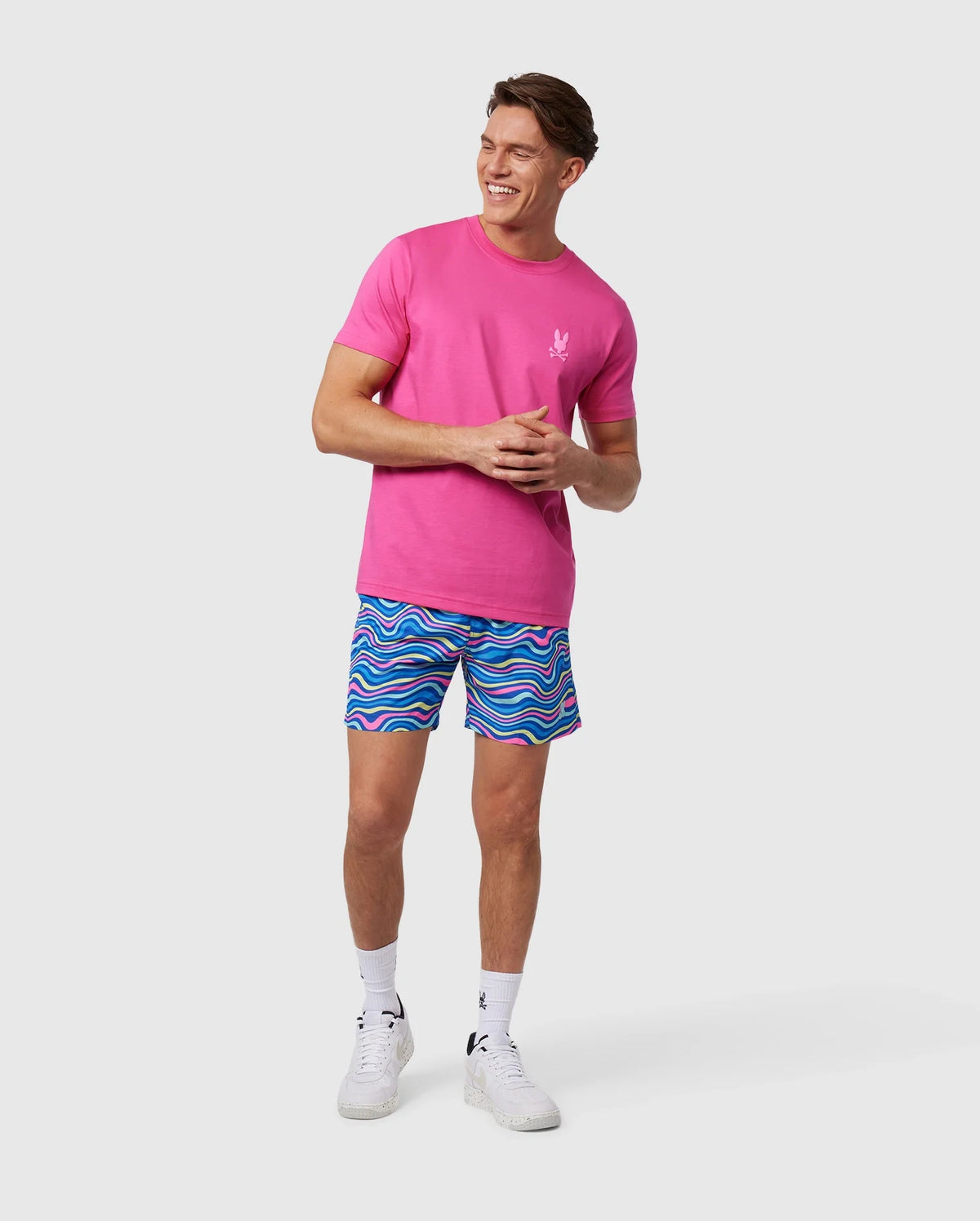 A man stands smiling, wearing a bright pink t-shirt with a small white logo on the chest and colorful quick-dry swim trunks, specifically the MENS CLARKSON LIGHTWEIGHT SWIM TRUNK - B6W120B2SW by Psycho Bunny, featuring a blue and pink wave-like pattern. He is also wearing white sneakers and socks, with a neutral background behind him.