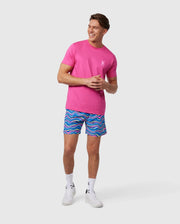 A man stands smiling, wearing a bright pink t-shirt with a small white logo on the chest and colorful quick-dry swim trunks, specifically the MENS CLARKSON LIGHTWEIGHT SWIM TRUNK - B6W120B2SW by Psycho Bunny, featuring a blue and pink wave-like pattern. He is also wearing white sneakers and socks, with a neutral background behind him.