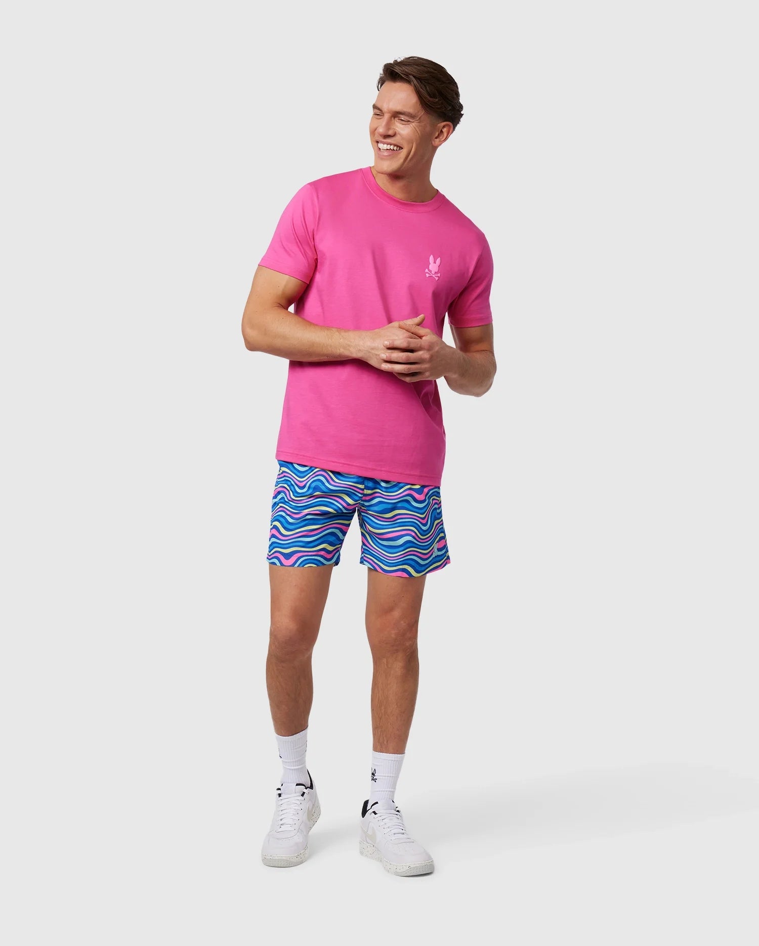 A man stands smiling, wearing a bright pink t-shirt with a small white logo on the chest and colorful quick-dry swim trunks, specifically the MENS CLARKSON LIGHTWEIGHT SWIM TRUNK - B6W120B2SW by Psycho Bunny, featuring a blue and pink wave-like pattern. He is also wearing white sneakers and socks, with a neutral background behind him.