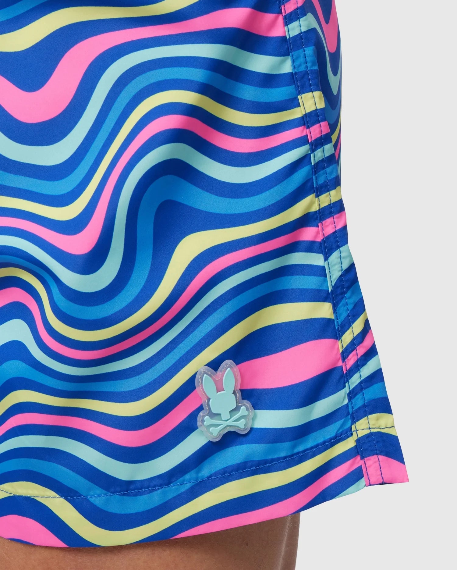 Close-up of colorful quick-dry swim trunks with blue, pink, and yellow wave-like stripes and a small bunny logo near the hem. The fabric, made from recycled polyester, features visible stitching lines. Product: MENS CLARKSON LIGHTWEIGHT SWIM TRUNK - B6W120B2SW by Psycho Bunny.