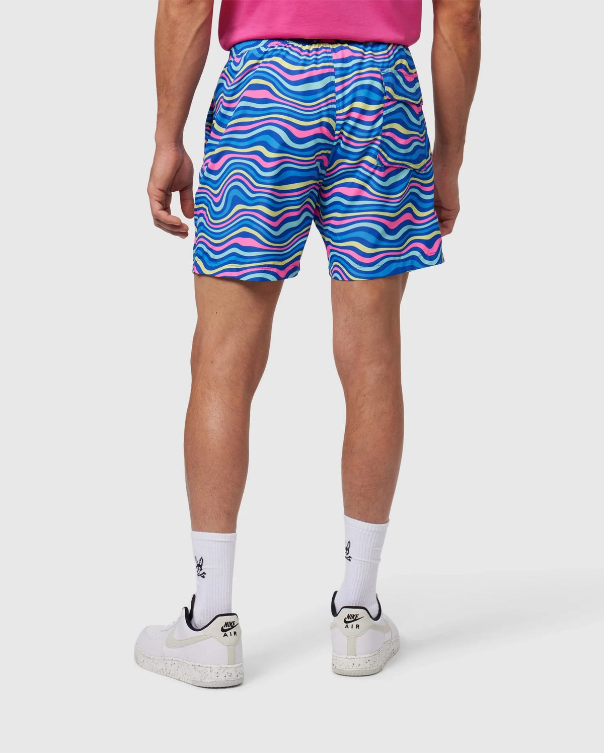 A person wearing brightly colored Psycho Bunny MENS CLARKSON LIGHTWEIGHT SWIM TRUNK - B6W120B2SW with a wave-like blue, pink, and white pattern. The individual is also sporting a pink shirt tucked into the shorts, white socks, and white sneakers. The image captures the back view of this stylish outfit made from recycled polyester.