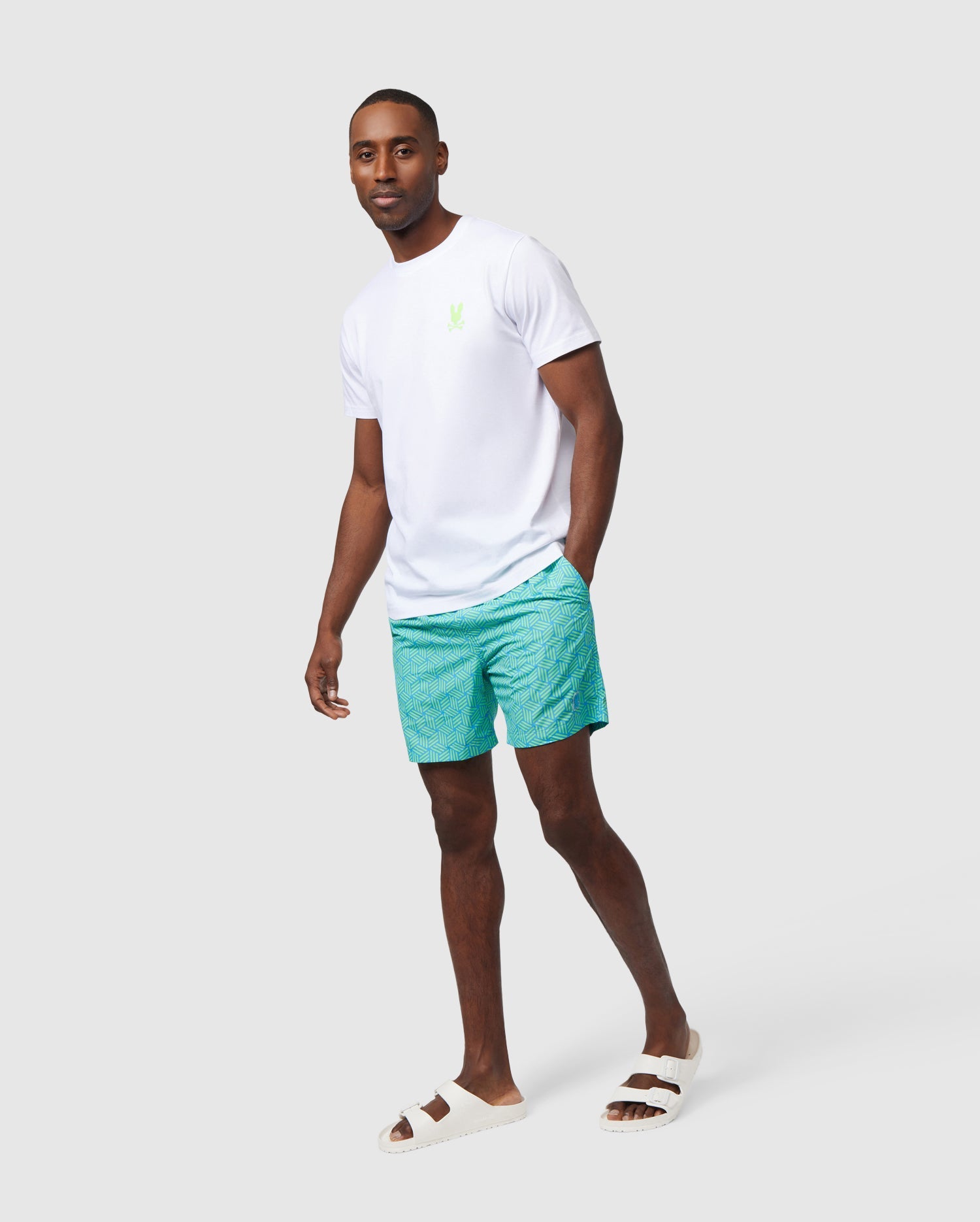 A man stands against a white background, wearing a white t-shirt with a green logo, Psycho Bunny's MENS JASPER LIGHTWEIGHT SWIM TRUNK in teal, and white slide sandals, looking directly at the camera.