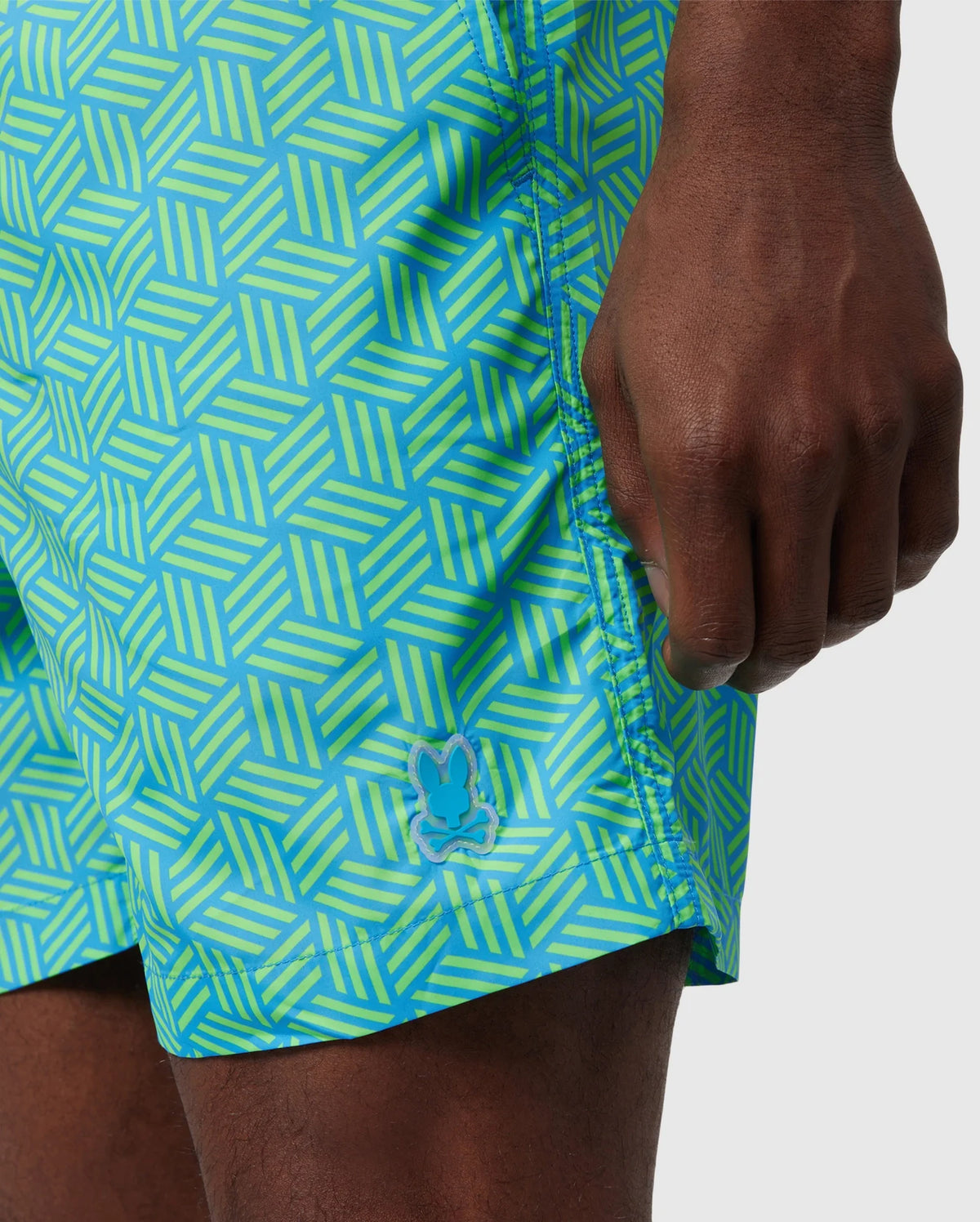 Close-up of a person's hand and part of their thigh, wearing a brightly patterned blue and green Psycho Bunny MENS JASPER LIGHTWEIGHT SWIM TRUNK - B6W168B2SW. A small logo is visible on the shorts.