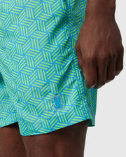 Close-up of a person's hand and part of their thigh, wearing a brightly patterned blue and green Psycho Bunny MENS JASPER LIGHTWEIGHT SWIM TRUNK - B6W168B2SW. A small logo is visible on the shorts.