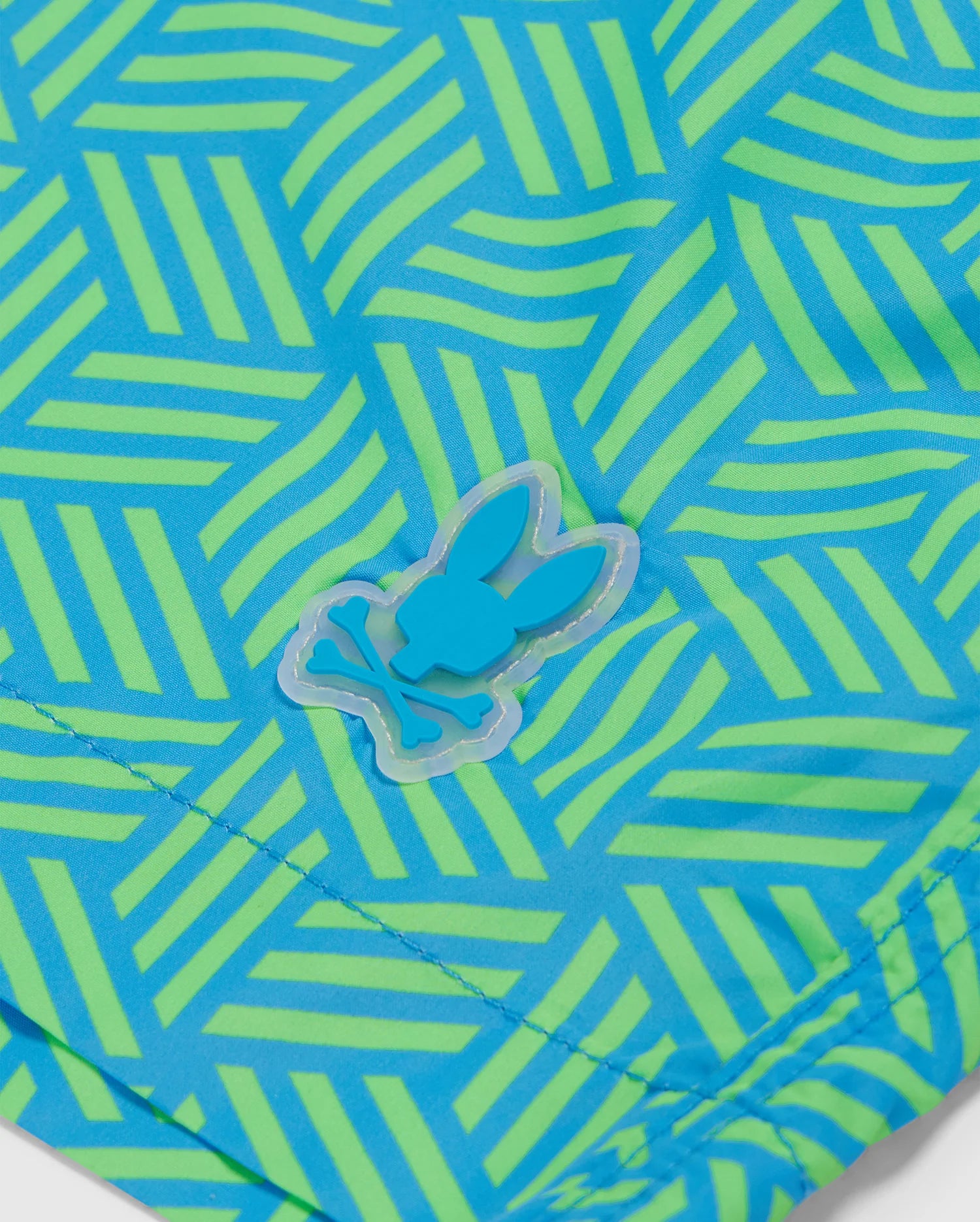 Close-up view of a Psycho Bunny MENS JASPER LIGHTWEIGHT SWIM TRUNK - B6W168B2SW in neon green and blue geometric-printed fabric with a blue rabbit-shaped sticker attached. The fabric texture is detailed with visible stitching.
