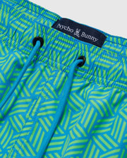 Close-up of vibrant green and blue geometric-printed swim shorts with a prominent "MENS JASPER LIGHTWEIGHT SWIM TRUNK - B6W168B2SW" label and blue drawstrings by Psycho Bunny.