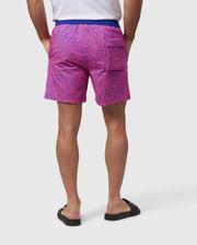 A person is standing with their back to the camera, wearing a white t-shirt and Psycho Bunny's VERONA PRINT SWIM TRUNK - B6W172B2SW with a wavy pattern made of quick-dry fabric. The black sandals complement the look, and their hands rest in the back pockets of the shorts. The background is plain white.