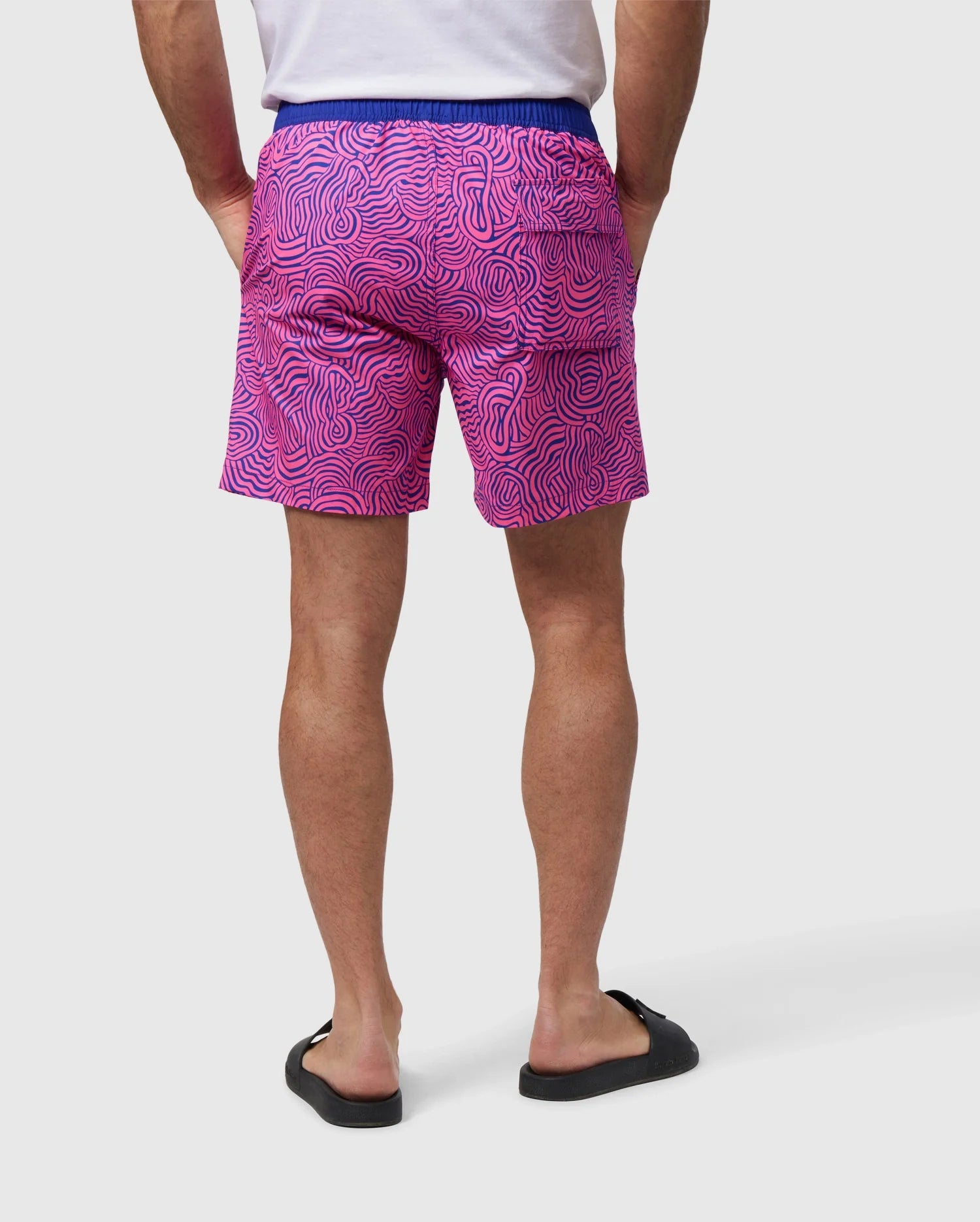 A person is standing with their back to the camera, wearing a white t-shirt and Psycho Bunny's VERONA PRINT SWIM TRUNK - B6W172B2SW with a wavy pattern made of quick-dry fabric. The black sandals complement the look, and their hands rest in the back pockets of the shorts. The background is plain white.